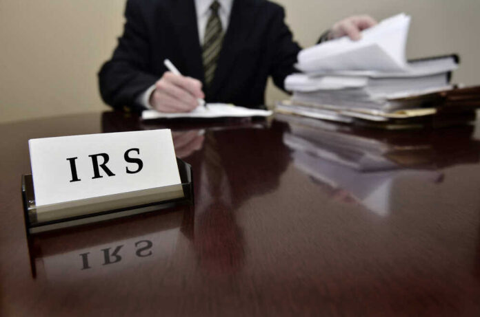 shutterstock_158874281.jpg featured image IRS sign on desk, person handling paperwork behind.