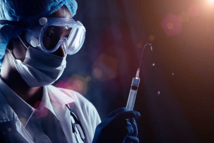 shutterstock_1880299123.jpg featured image Person in PPE holding syringe with liquid droplets.