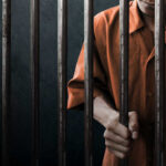 Man in orange prison uniform behind jail bars.
