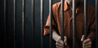 Man in orange prison uniform behind jail bars.