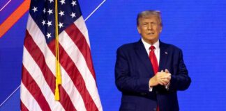 Donald Trump in suit near the American flag on stage.