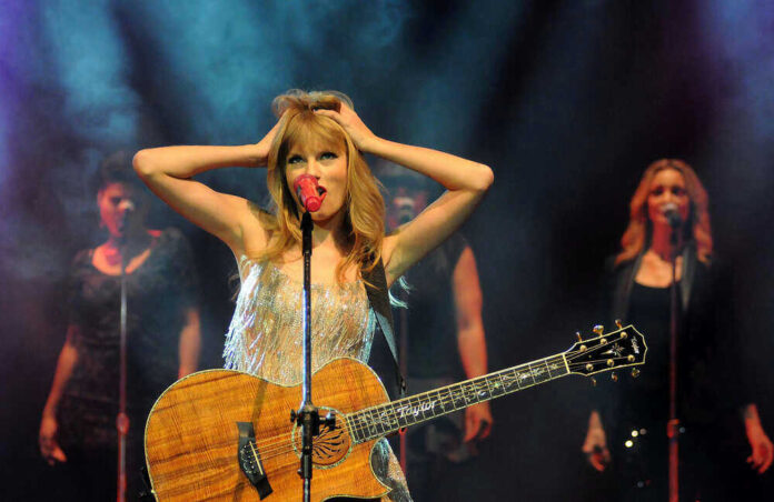 Taylor Swift holding guitar on stage, background singers behind