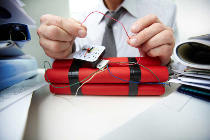 A person in a shirt and tie is holding wires connected to a timer on a red explosive device.
