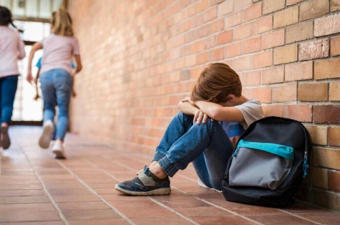 shutterstock_1136889179.jpg Sad child sitting against brick wall, kids running away.