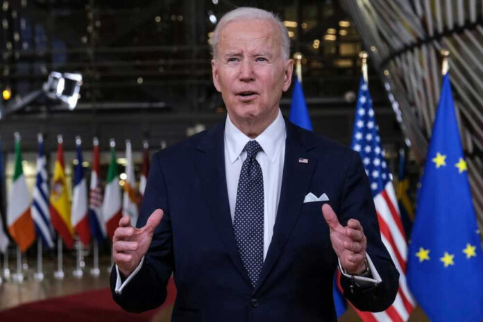 Joe Biden in suit speaking, international flags in background.