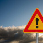 Mobs Storm US Consulate – Many Dead! A triangular warning sign with an exclamation mark against a cloudy sky
