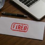 An envelope marked 'FIRED' on a wooden desk next to a clock and a laptop