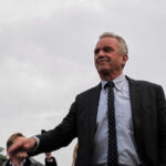 RFK Jr. Ties Federal Funding To Hospital Meals Robert F. Kennedy Jr. in a suit stands outdoors, cloudy sky background.
