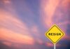 A yellow warning sign with the word 'RESIGN' against a colorful sunset sky