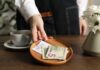 A hand placing cash and a receipt on a wooden plate next to a cup of coffee