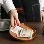 A hand placing cash and a receipt on a wooden plate next to a cup of coffee