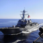 Pentagon Boat War Escalates Warship with American flag sailing in the ocean.