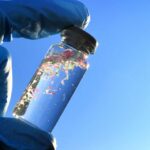 A gloved hand holding a glass vial containing colorful microplastics against a blue sky