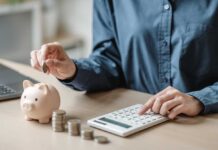 Savings Crash Near Pandemic Lows Person placing a coin into a piggy bank while using a calculator