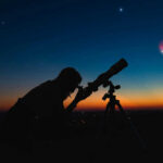 Tiny Planet Exposes Cosmic Carve-Out Silhouette of person using telescope against colorful twilight sky.