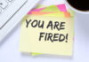 A sticky note with the message 'YOU ARE FIRED!' placed on a desk next to a keyboard and a cup of coffee