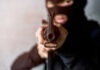Person in balaclava aiming rifle at the camera.