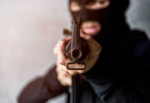 Masked Gunman Ambushes Kids’ Cookout – 8 Shot Person in balaclava aiming rifle at the camera.