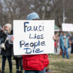 Shredded Emails Rock Fauci’s Inner Circle Person holding sign reading Fauci Lies, People Die.