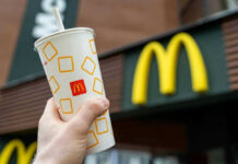 McDonald’s ‘Heist’ Menu Lands – Blink And Miss It Hand holding McDonalds cup in front of restaurant sign.