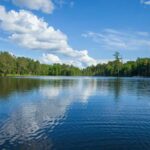 Lake Day Turns Deadly — Chilling Mystery A tranquil lake surrounded by trees under a blue sky with fluffy clouds