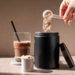 Hand pouring protein powder into a container next to a glass of iced coffee