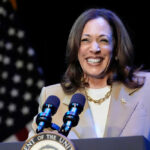 Kamala’s Speech Gets Weird – What’s She Hiding? Kamala Harris smiling at a podium with microphones